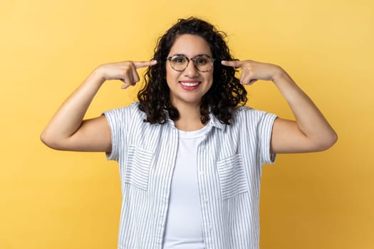Portrait of smiling friendly positive woman with dark wavy hair in eyeglasses, standing pointing at her new spectacles, looking at camera. Indoor studio shot isolated on yellow background.