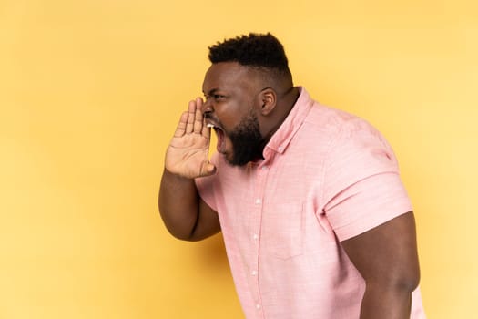 Side view of angry aggressive man wearing pink shirt standing, holding arms near wide open mouth and screaming, trying to get attention. Indoor studio shot isolated on yellow background.