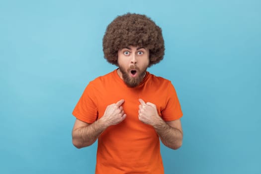 Portrait of man with Afro hairstyle in orange T-shirt points himself, looking with surprised expression, shocked and proud of win, unbelievable success. Indoor studio shot isolated on blue background.