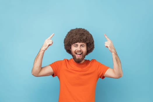 Portrait of extremely happy man with Afro hairstyle in orange T-shirt pointing finger up with toothy smile on face, clever smart man has idea, start up. Indoor studio shot isolated on blue background.