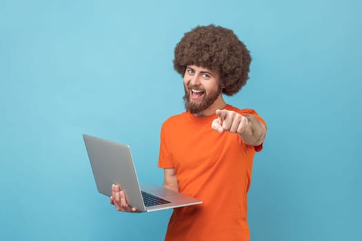 Portrait of happy man with Afro hairstyle wearing orange T-shirt standing with portable computer in hand, looking and pointing at camera. Indoor studio shot isolated on blue background.