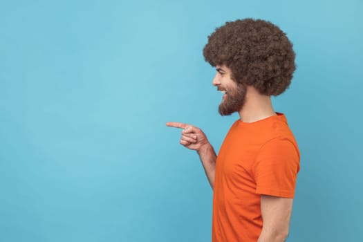Side view of man with Afro hairstyle wearing orange T-shirt pointing finger away with toothy smile on face, empty space for your advertisement. Indoor studio shot isolated on blue background.