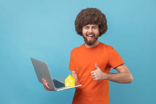Smiling man with Afro hairstyle wearing orange T-shirt standing with laptop and paper house, showing thumb up, like gesture, helping with rent. Indoor studio shot isolated on blue background.