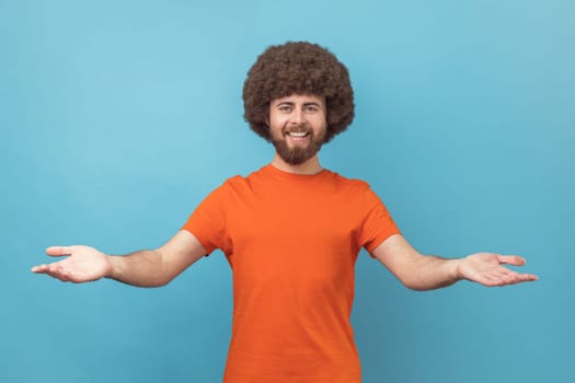 Man with Afro hairstyle wearing orange T-shirt sharing opening hands looking at camera with kind smile, greeting and regaling, happy glad to see you. Indoor studio shot isolated on blue background.