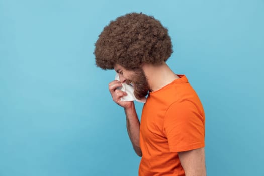 Side view of depressed man with Afro hairstyle wearing orange T-shirt holding head down, hiding his face in hand and crying, feeling desperate. Indoor studio shot isolated on blue background.