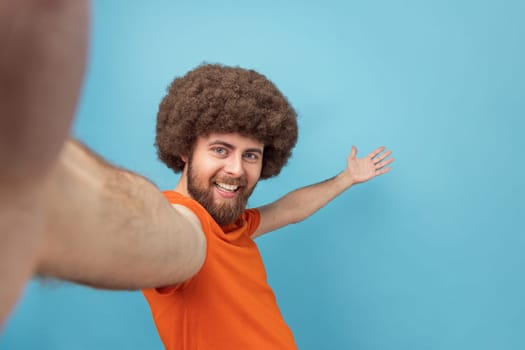 Portrait of king pleased man with Afro hairstyle broadcasting livestream, talking to followers, inviting friends, showing welcome gesture, POV. Indoor studio shot isolated on blue background.