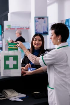 Drugstore worker putting vitamin in customer shopping bag at medical retail store counter desk. Smiling buyer standing near pharmaceutical assistant at checkout and buying dietary supplements