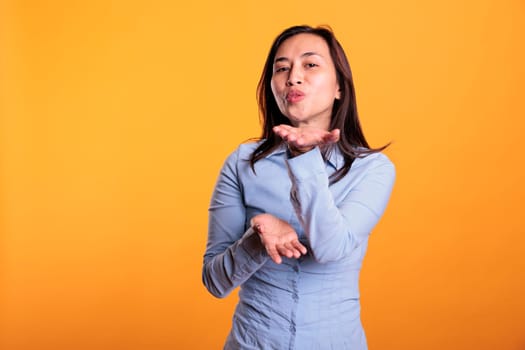 Confident joyful filipino woman blowing air kissed during studio shot, posing over yellow background. Attractive model expressing love, looking at the camera with a flirty smile. Romantic gesture