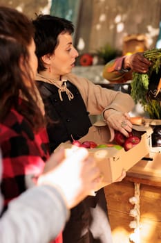 Diverse farmers selling natural fruits and veggies in boxes, serving senior man and young woman at farmers market. Smiling people looking at healthy organic colorful products.