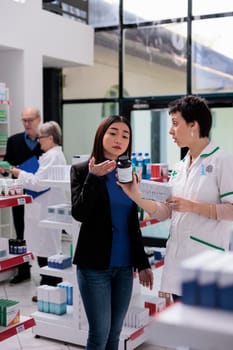 Young asian woman returning wrong vitamin package in drugstore, asking pharmaceutical consultant to exchange dietary supplements box. Customer showing chemist multivitamin components on bottle