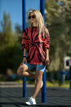 blonde athletic girl stands on the sports ground in a t-shirt, breeches, white sneakers and white headphones