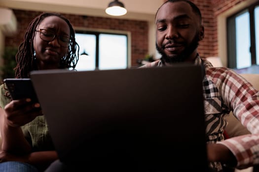 Woman browsing social media on smartphone while boyfriend is watching online series on laptop while sitting on couch. African american couple spending free time together looking at video content.