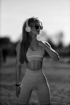 blonde athletic girl stands on the football field in a tank top, breeches, white sneakers and white headphones black and white