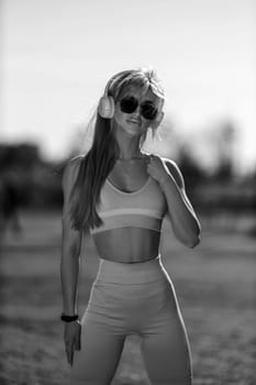 blonde athletic girl stands on the football field in a tank top, breeches, white sneakers and white headphones black and white