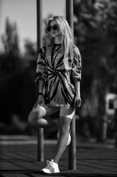 blonde athletic girl stands on the sports ground in a t-shirt, breeches, white sneakers and white headphones black and white