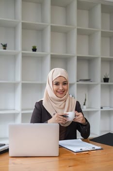 Asian Muslim business woman holding a coffee mug siting in the modern office. business people, diversity and office concept.