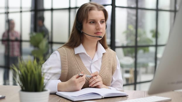 Attractive confident business woman, office manager, wearing headset using laptop while making, writing financial report, using pen, on paper working indoors.