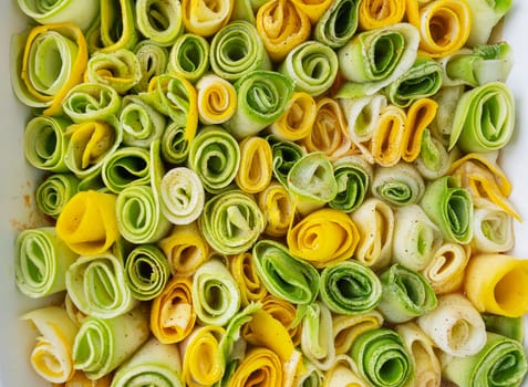 Small rolls of zucchini green and yellow with creamy sauce in a white baking dish against the background of a checkered napkin. Top view, close-up