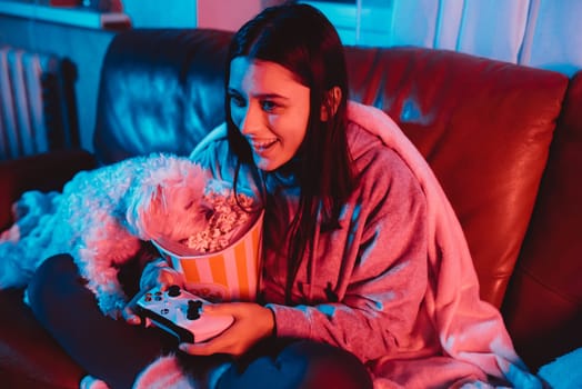 A gamer or a streamer girl at home in a dark room with a game controller and popcorn bucket playing with her dog and sits in front of a monitor or TV. High quality photo