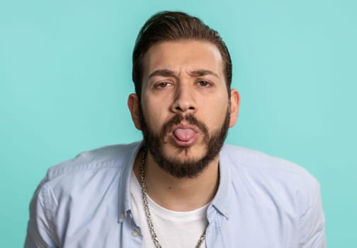 Comical funny young lebanese man making playful silly facial expressions and grimacing, fooling around, showing tongue. Handsome arabian crazy guy bullying isolated on studio blue background indoors