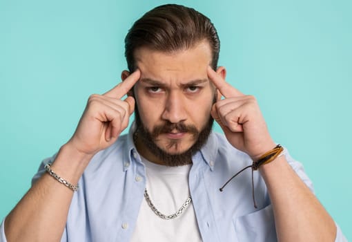 Handsome lebanese bearded man in shirt rubbing temples to cure headache problem, suffering from tension, migaine, stress, grimacing in pain, high blood pressure isolated alone on blue background