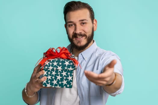 Positive smiling young lebanese man presenting birthday gift box stretches out hands, offer wrapped present career bonus, celebrating party sale. Bearded arabian guy isolated on blue studio background