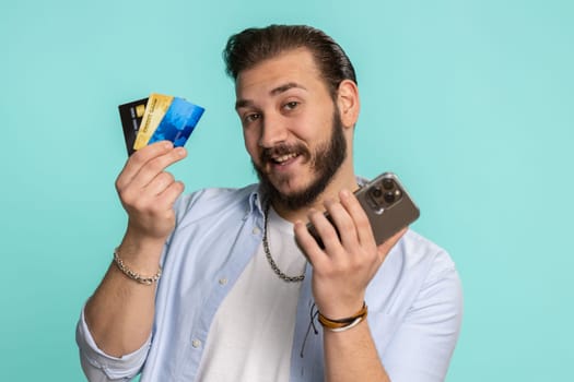 Sincere lebanese man customer using credit bank cards and smartphone while transferring money, purchases online shopping, payment. Finance and internet. Handsome guy isolated alone on blue background