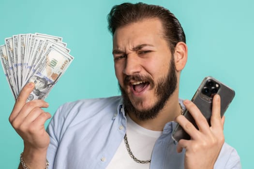 Lebanese man looking smartphone display sincerely rejoicing win, receiving money dollar cash banknotes, success lottery jackpot luck, income salary wage. Arabian guy isolated on studio blue background