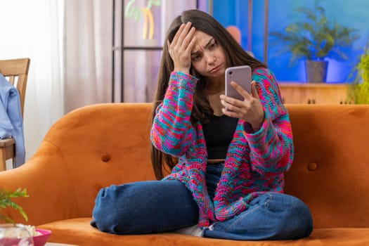 Sad displeased woman use smartphone typing browsing, loses becoming surprised sudden lottery results, bad news, fortune loss, fail, deadline, virus. Young Caucasian girl at home living room on couch