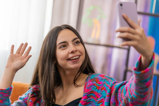 Happy woman blogger taking selfie on smartphone, communicating video call online with subscribers, recording stories for social media. Portrait of Caucasian girl at home apartment living room on sofa