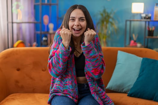 Happy Caucasian woman shouting, celebrating success, winning, goal achievement good news, lottery luck, victory, watching TV championship at modern home apartment. Girl in living room sitting on couch