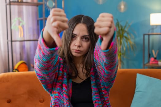 Dislike. Upset young woman showing thumbs down sign gesture, expressing discontent, disapproval dissatisfied bad work at modern home apartment indoors. Displeased Caucasian girl in living room on sofa