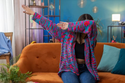 Trendy cheerful positive young woman having fun dancing and moving to rhythm, dabbing raising hands, making dub dance gesture. Portrait of Caucasian girl celebrate win at home apartment room on sofa
