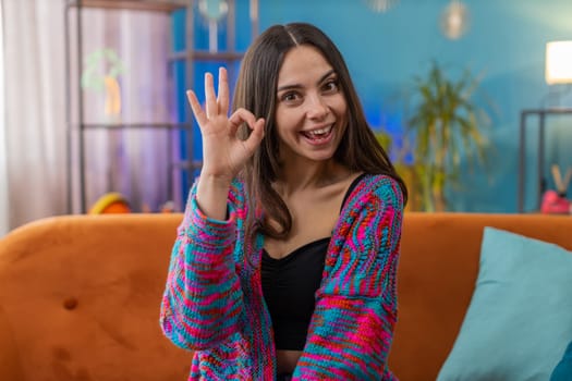 Okay. Happy cheerful young woman looking approvingly at camera showing ok gesture, positive like sign, approve something good. Portrait of Caucasian girl at home modern apartment room on orange sofa