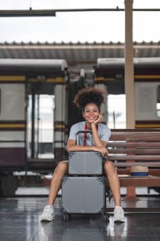 Asian African female tourist traveler holding mobile phone smart phone sitting at train station. Confident smiling teenager girl playing smart phone laptop computer on suit case at station