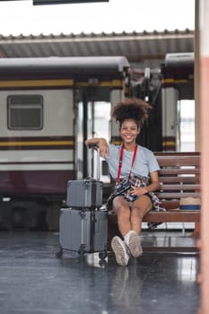 Asian African female tourist traveler holding mobile phone smart phone sitting at train station. Confident smiling teenager girl playing smart phone laptop computer on suit case at station