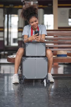 Asian African female tourist traveler holding mobile phone smart phone sitting at train station. Confident smiling teenager girl playing smart phone laptop computer on suit case at station