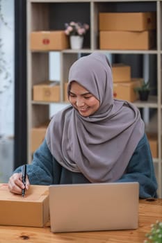Muslim asian woman freelancer working and checklist and writing order with cardboard box at home - SME business online and delivery concept..