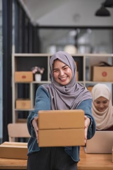 A happy young Asian Muslim female online seller working with her employee in the office. SME business, online shop owner..