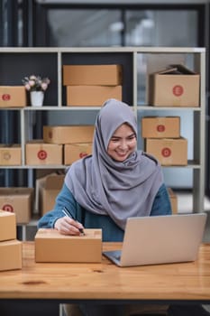 Muslim asian woman freelancer working and checklist and writing order with cardboard box at home - SME business online and delivery concept..