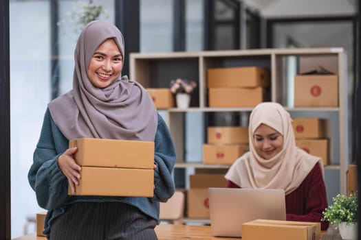 A happy young Asian Muslim female online seller working with her employee in the office. SME business, online shop owner..