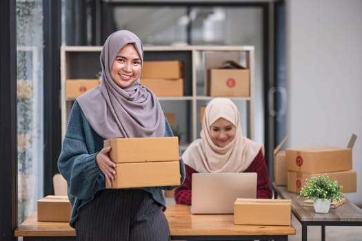 A happy young Asian Muslim female online seller working with her employee in the office. SME business, online shop owner..
