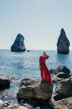 Woman travel sea. Young Happy woman in a long red dress posing on a beach near the sea on background of volcanic rocks, like in Iceland, sharing travel adventure journey