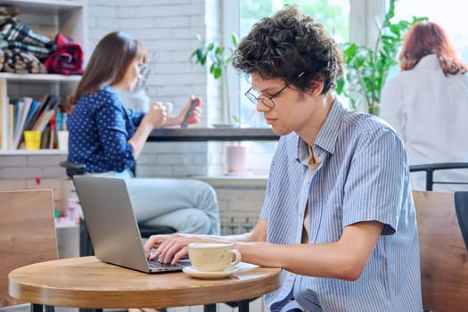 Handsome young curly guy college student typing on laptop sitting at table at coffee shop with cup of coffee. Internet online technology for leisure communication learning, chat, blog, youth lifestyle