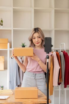 Asian beautiful woman sell clothes online live streaming at home. Young female use mobile phone video call shows goods to customer and present detail. Remote buying and purchase shopping concept.