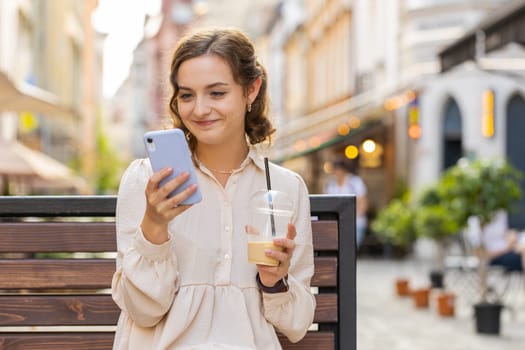 Smiling pretty young woman using smartphone typing text answering messages chatting online looking mobile screen social media app. Girl tourist sits on bench on city street drinks cold coffee with ice