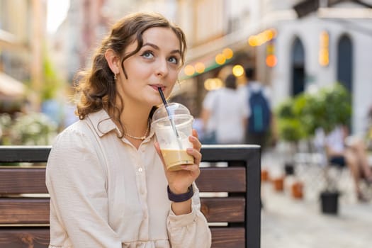 Happy young woman enjoying morning cold coffee drink with ice and smiling outdoors. Relaxing, taking a break. Girl sits on bench on city sunny street, drinking coffee to go. Town lifestyles outside