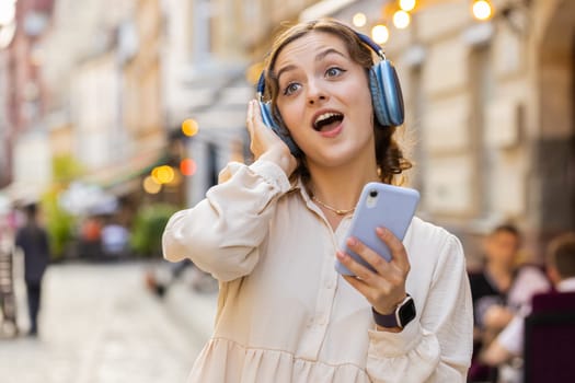 Happy relaxed overjoyed pretty young woman in wireless headphones choosing, listening favorite energetic disco rock n roll music dancing outdoors. Girl walking in urban city sunshine street background