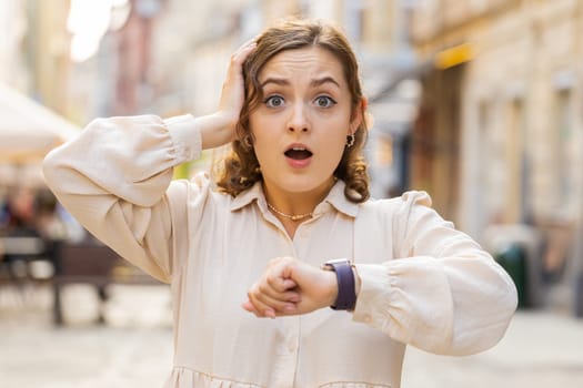 Scared woman worrying to be punctual, with anxiety checking time on watch, running late to work or transport, being in delay, deadline outdoors. Girl tourist walking in city street. Town lifestyles
