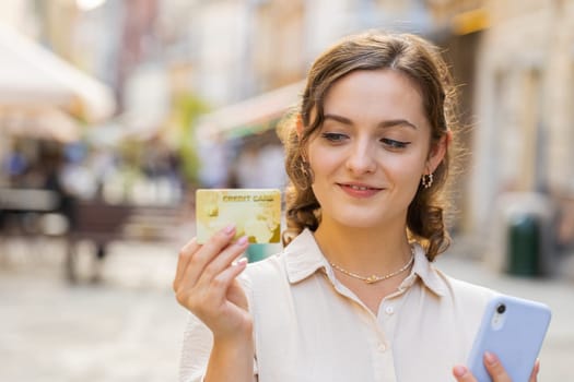 Young woman using credit bank card smartphone while transferring money, purchases online shopping, order food delivery, booking hotel room. Lovely girl walking in urban city sunny street outdoors